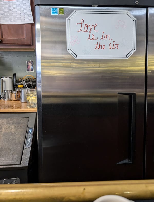 Stainless steel restaurant refrigerator with a dry erase board with the words "Love is in the air" on it.